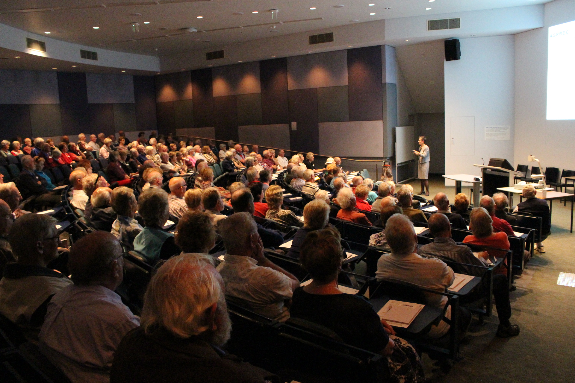 Older adults fill a lecture theatre, listening to a suited woman explain aspects of the ASPREE trial displayed on the large screen behind her