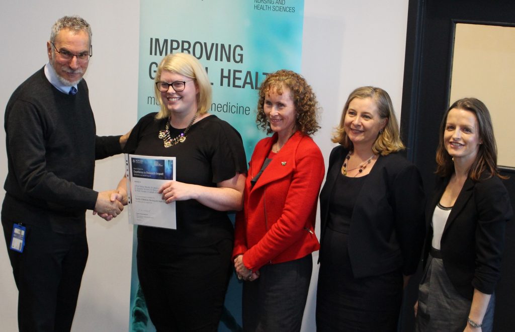Woman with blond hair accepts a certificate and shakes the presenter's hand. Standing alongside are three women, all senior managers working on the ASPREE trial.