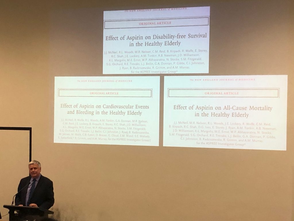 A grey haired man stands at a lectern with images of three ASPREE papers published in the New England Journal of Medicine projected on the screen behind him.