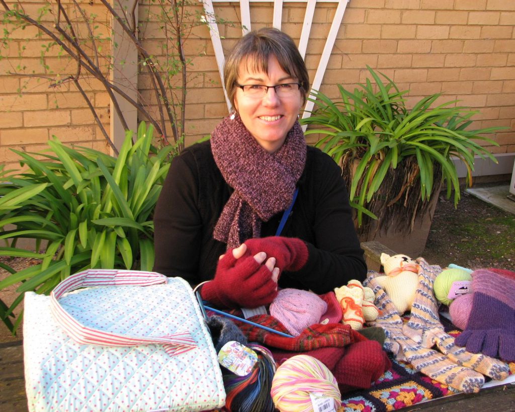 White woman with short dark hair and glasses sits at a table covered with hand knitted articles, including gloves and socks