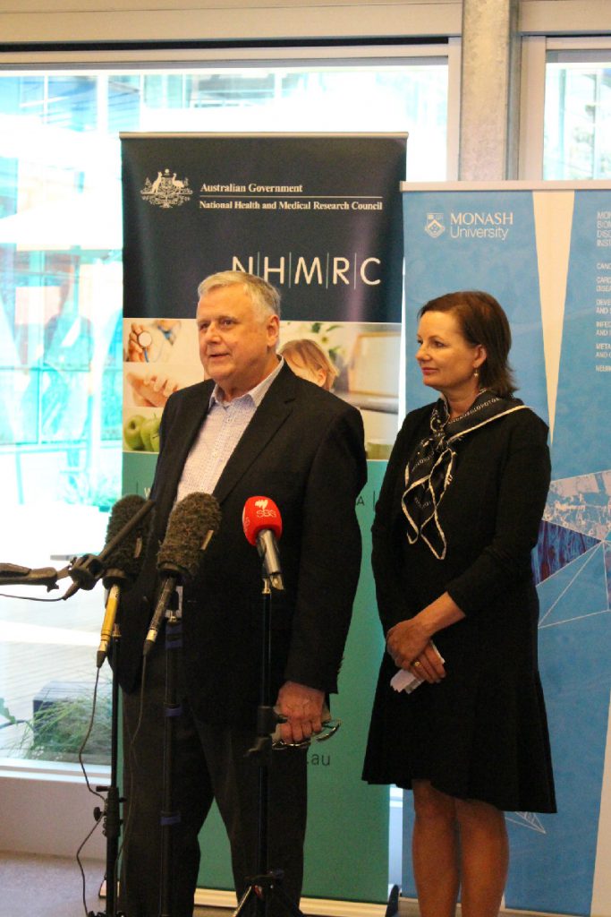 Grey haired, man in a suit is talking in front of TV microphones next to a women dressed in a dark suit. They stand in front of NHMRC and Monash University banners