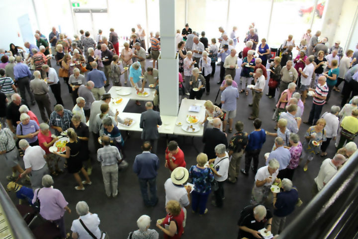 Looking down onto a large group of older adults mingling with a cuppa and food. 