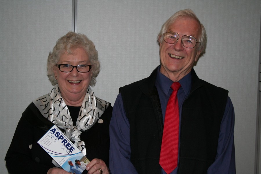 Older lady with black rimmed glasses holds an ASPREE brochure, next to an older gentleman wearing a red tie and blue shirt.