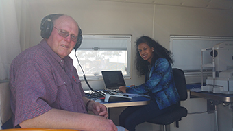 An older gent wearing a set of black headphones sits adjacent to a young woman with long dark hair in a blue jacket inside an ASPREE retcam van. The woman sits in front of audiometry equipment.