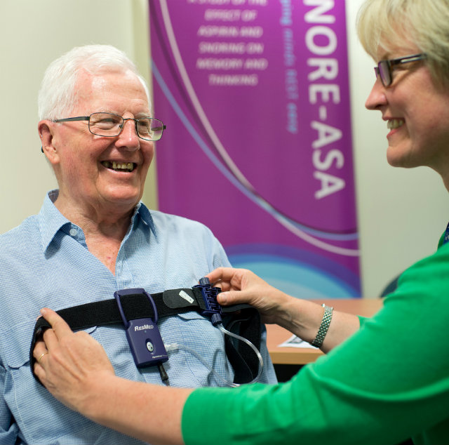 An older gentleman in a blue shirt has the SNORE-ASA  portable sleep apnoea device placed across his chest by a researcher