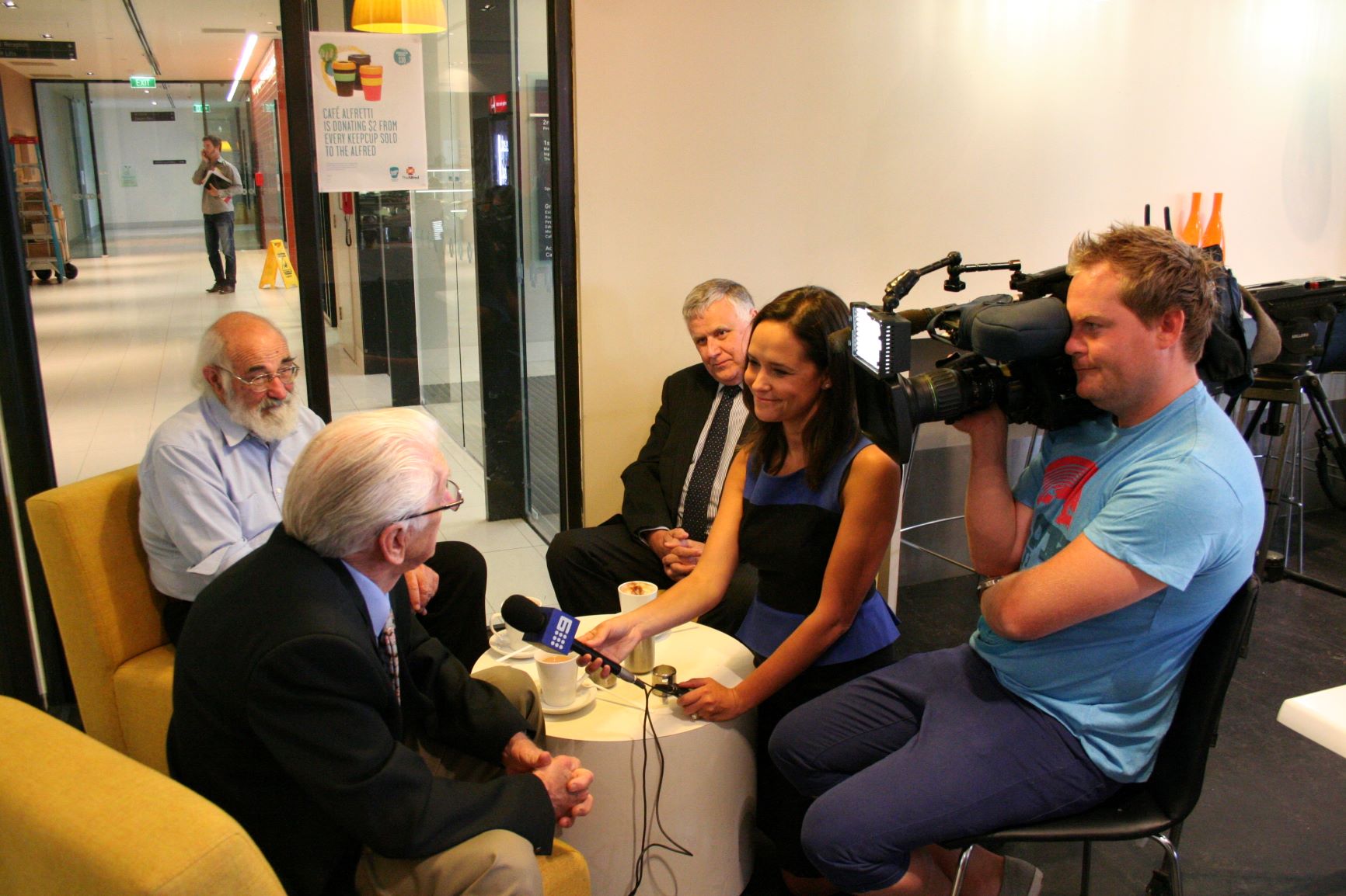 Older grey haired gent being interviewed by a news crew inside a cafe. 