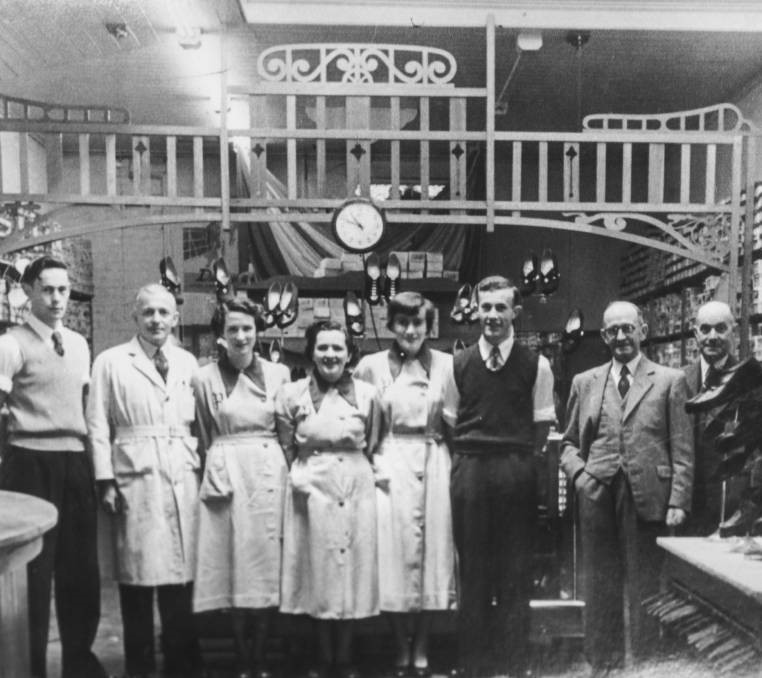 A black and white photograph of men and women with shelves of shoes and a clock face behind them