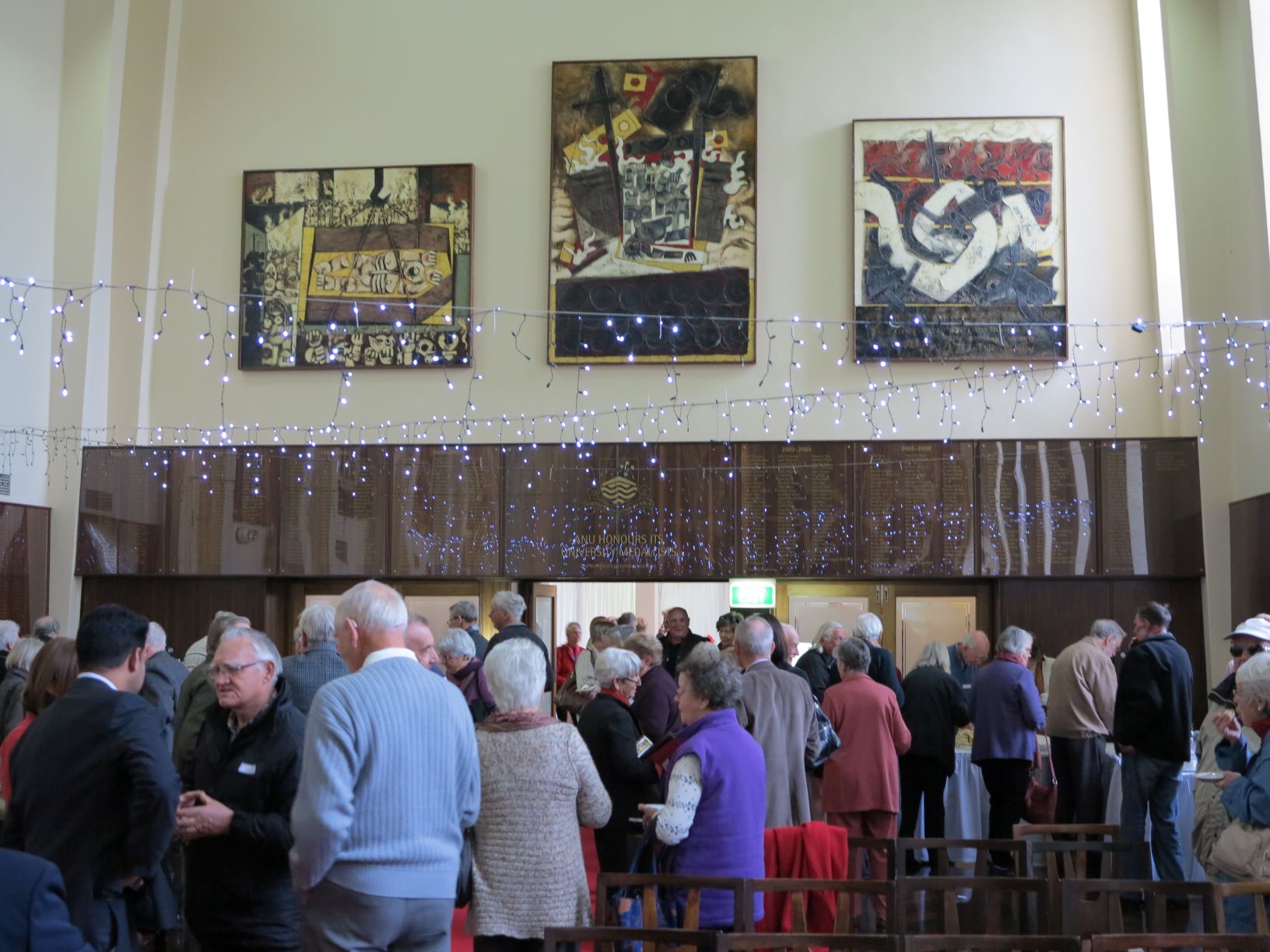 Group of older Australians mingling and talking in a large hall with three abstract art works placed high on the wall.