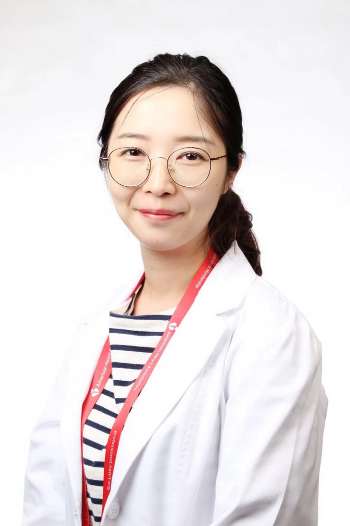 Woman with dark hair, glasses in a white coat, in front of a white background, smiles at the camera