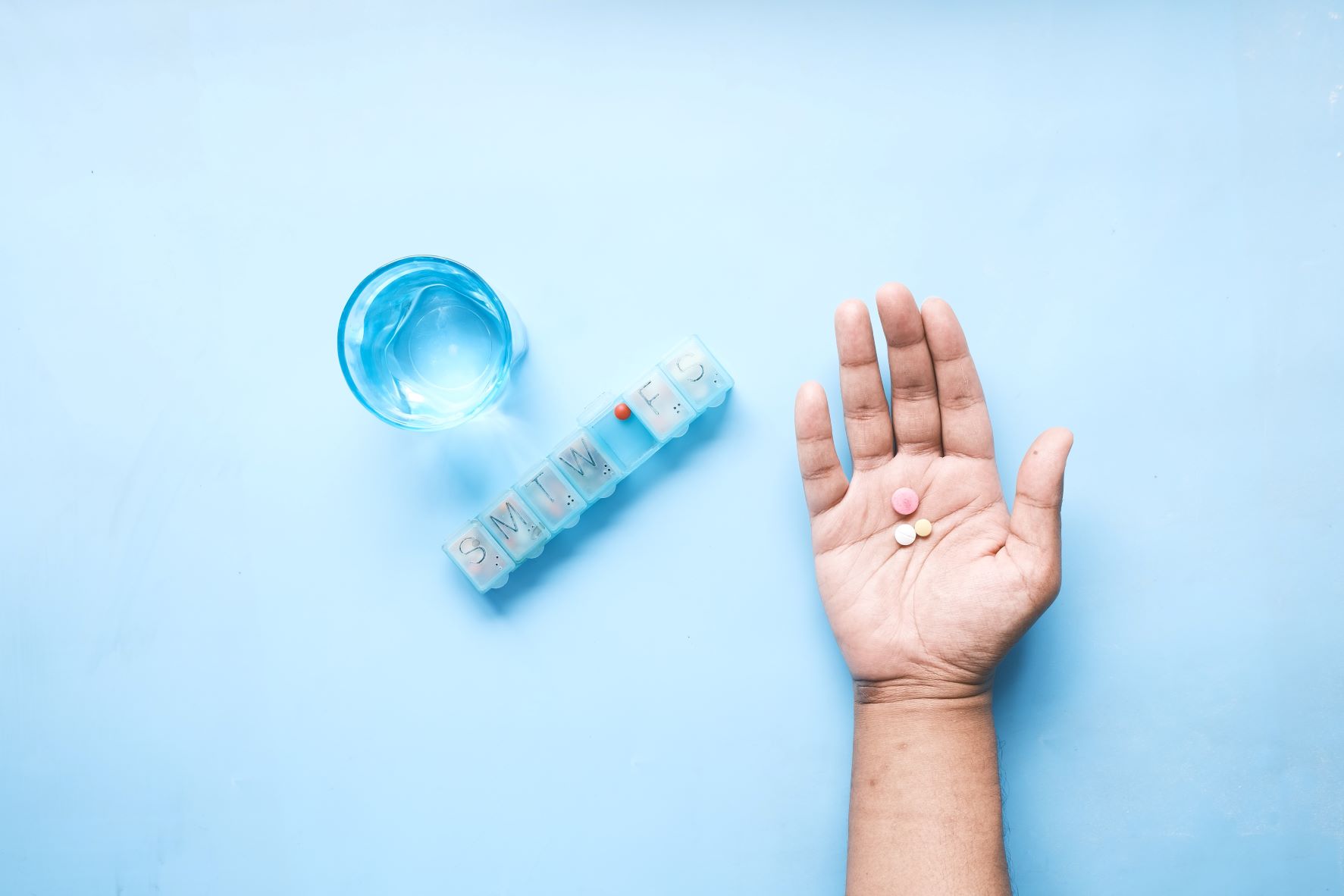 Glass water, pill box and an open hand with 3 tablets laid on the palm, in front of blue background